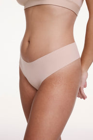 On a white studio backdrop, Sonnie wears the No-Show Low Rise Thong in Beige, shown at an angle to highlight the low-rise fit and smooth, elastic-free edges designed to stay invisible under clothing.