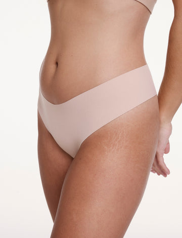 On a white studio backdrop, Sonnie wears the No-Show Low Rise Thong in Beige, shown at an angle to highlight the low-rise fit and smooth, elastic-free edges designed to stay invisible under clothing.