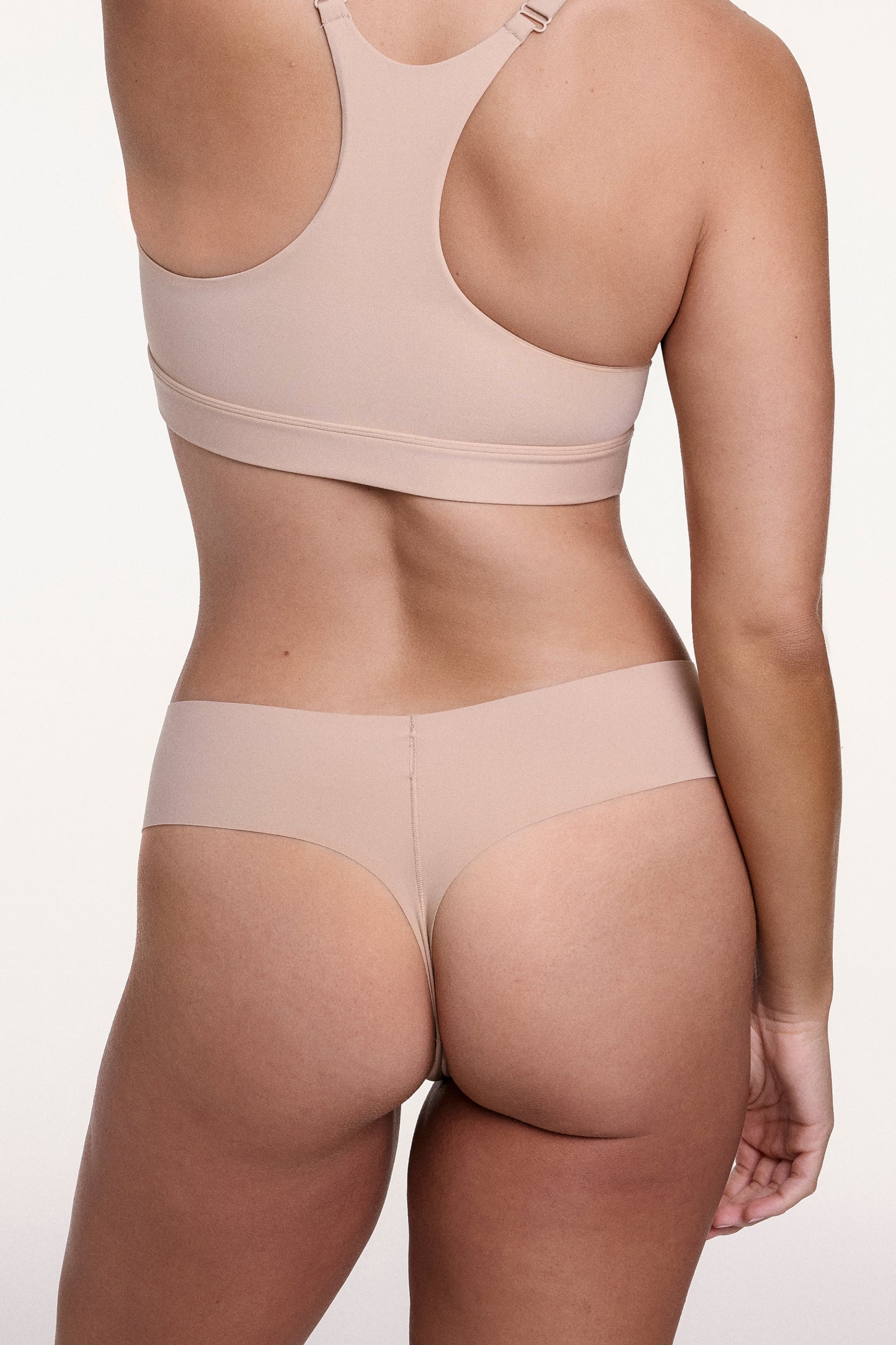 On a white studio backdrop, Sonnie wears the No-Show Low Rise Thong in Beige, shown from the back with minimal coverage and a smooth, raw-edge finish designed to lay flat under clothing.