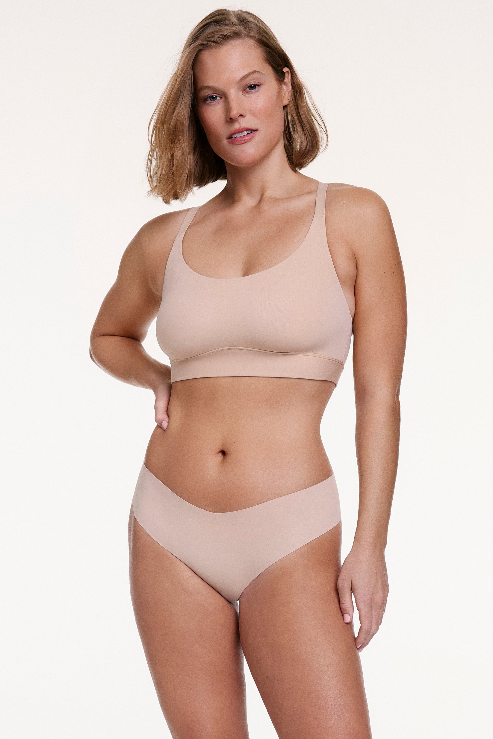 On a white studio backdrop, Sonnie wears the No-Show Low Rise Thong in Beige with a matching bra, showing the low-rise fit and soft, no-show fabric for a smooth, second-skin feel.