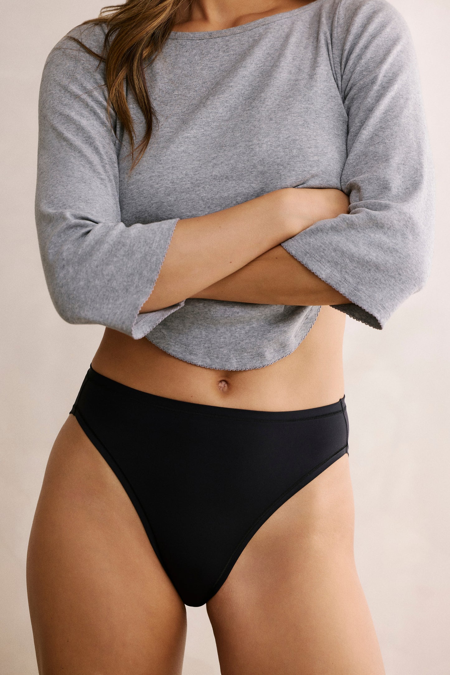In a softly lit room, a model wears the Bliss French Cut underwear in Black, standing with a gray top slightly lifted. The high-cut leg and smooth microfiber fabric are visible, with a no-show waistband designed to sit flat against the skin. sizing: Darion wears size Small