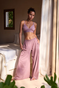 In a sunlit bedroom, a model wears the Bliss Triangle Bralette in Mirage with matching underwear and wide-leg lounge pants, standing beside a bed near an open window. The deep V neckline and adjustable straps are visible, with soft microfiber fabric and a wireless band providing comfortable lift.