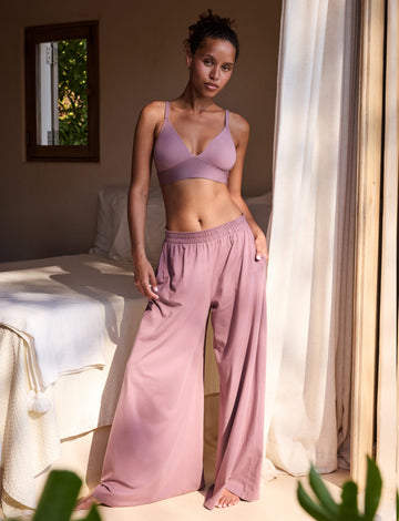 In a sunlit bedroom, a model wears the Bliss Triangle Bralette in Mirage with matching underwear and wide-leg lounge pants, standing beside a bed near an open window. The deep V neckline and adjustable straps are visible, with soft microfiber fabric and a wireless band providing comfortable lift.