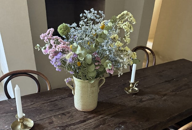 Lifestyle mood shot of a wildflower arrangement in a ceramic vase on a dark wood dining table with a brass candlestick