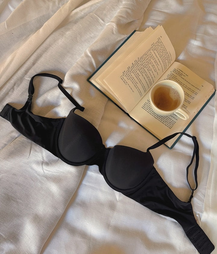Flat lay of Base Strapless Bra in Black and ComfortMesh Highwaist Brief in Black on white linen sheets beside an open book and cup of coffee