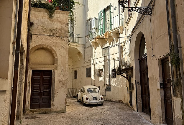Lifestyle mood shot of a vintage cream VW Beetle parked in a sunlit stone European courtyard with arched doorways and balconies