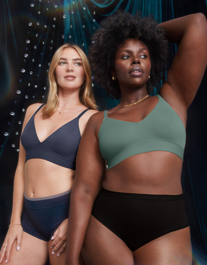 Megan (left) wearing the Bliss Triangle Bralette in Navy and Akesha (right) wearing the Bliss Triangle Bralette in Rainforest. They stand confidently in front of a dark, draped background with crystal accents, illuminated by soft lighting. Akesha poses with her arm raised behind her head.