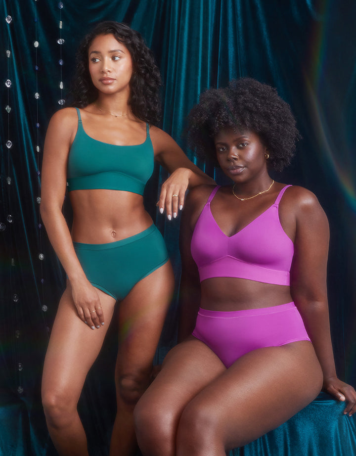 Ally in the Bliss Scoop and Highwaist Brief in Everglade stands next to Akesha, who is wearing the Bliss Triangle and Highwaist Brief in Berry. Ally rests her arm on Akesha’s shoulder, both posing confidently against a deep blue-green velvet backdrop adorned with crystal accents.