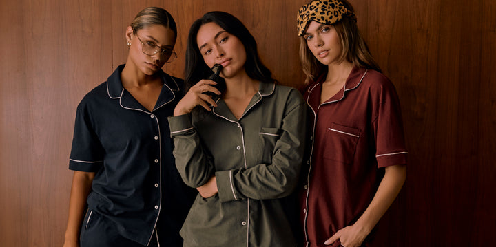 Three models pose against a wood-paneled wall wearing Harper Wilde’s Pima Pajama Sets. From left to right: one wears Navy with glasses, one wears Olive holding a sleep mask to her lips, and one wears Bordeaux paired with a leopard-print sleep mask. All tops  featuring white piping and button-front details.