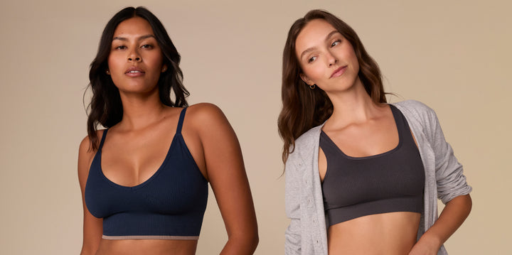 Two models stand casually against a beige background. The left model is wearing the Lounge Rib Triangle Bralette in True Blue and the right model is wearing the Lounge Scoop Bralette in Charcoal under a open gray cardigan.