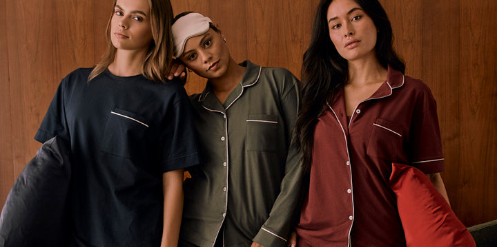 Three models wearing Harper Wilde Pima Pajamas in navy, olive, and bordeaux. The pajamas feature soft cotton fabric with contrast piping and chest pockets, styled for comfort and relaxation. The center model wears a matching eye mask and rests her head on the model beside her.