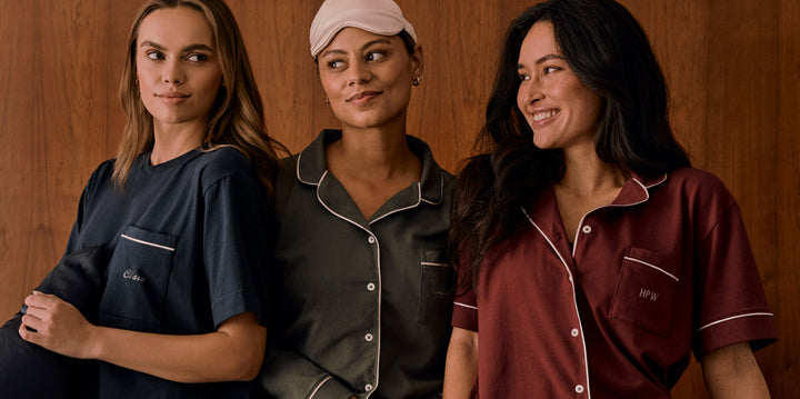 Three models wearing the Perfect Pima Pajama tops stand in front of a warm wood backdrop. From left to right: the Navy top with "Clara" embroidered on the chest pocket, the Olive top with no visible embroidery, and the Bordeaux top with "HPW" embroidered on the pocket. Each top features white piping and a soft, relaxed fit.