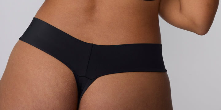 On a clean studio backdrop, three models are shown wearing black no-show underwear styled with matching bras. The image highlights smooth, elastic-free waistbands and minimal coverage silhouettes designed to sit flat under clothing.