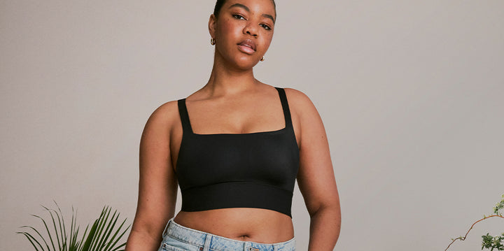 Zarria wears the Bliss Square Neck Bralette in Black, paired with light-wash jeans. She stands confidently against a neutral backdrop with soft lighting and minimal greenery.