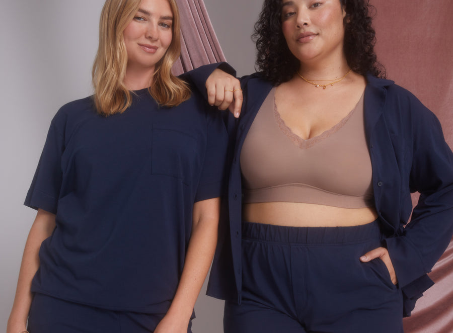 Two models dressed in Harper Wilde Perfect Pima Pajamas. One wears a Pima T-Shirt and Pant set in Navy while the other wears a Bliss Lace Bralette in Muave with Navy Pima shorts, standing against a pink backdrop.