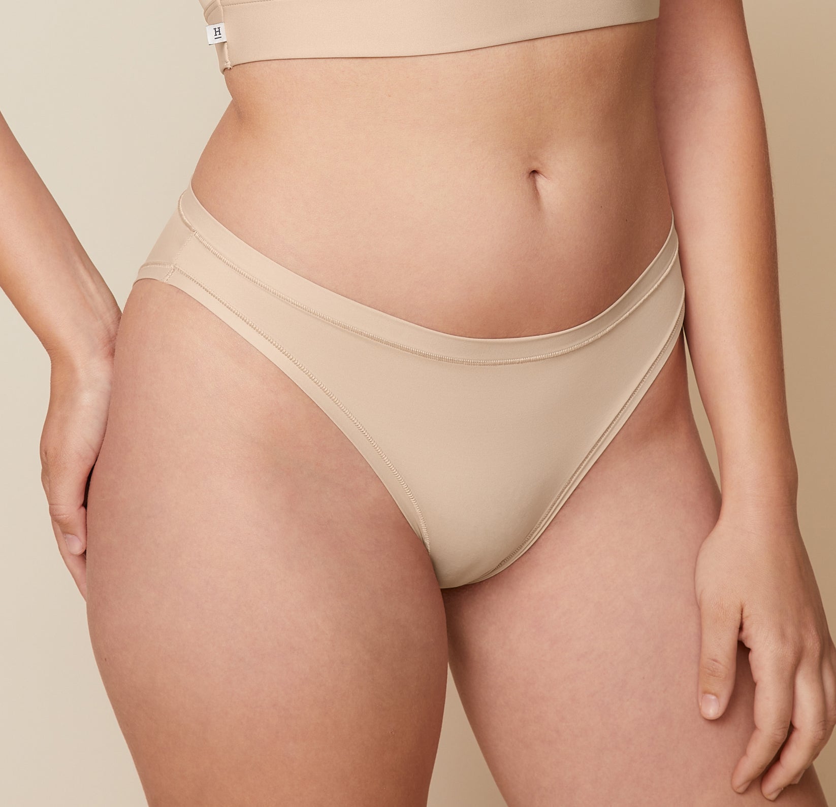 A close-up side view of Carli, focusing on her lower body. She is wearing the Bliss Bikini in Beige, showcasing the design and fit of the underwear. 