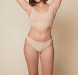 Carli is standing facing forward with her arms raised above her head, smiling. She is wearing the Bliss Racerback Bralette and the Bliss Bikini in Beige, showcasing the set from the front. sizing: Carli wears size Medium