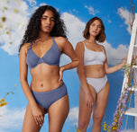 Naisha, on the left, poses confidently wearing the Base Unlined Bra and Bliss Hiphugger in the color Twilight. Carli, on the right, stands beside her, wearing the Bliss Scoop Bralette and Bliss Bikini in the color Tidal. Both models are set against a vibrant sky backdrop with delicate flowers in the foreground, highlighting the comfort and style of the products.