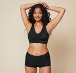 Naisha smiles with arms raised, wearing the Bliss Sleek Lined Bralette and Bliss Boyshort in Black. The bralette features a seamless design and V-neckline, paired with the comfortable boyshorts.