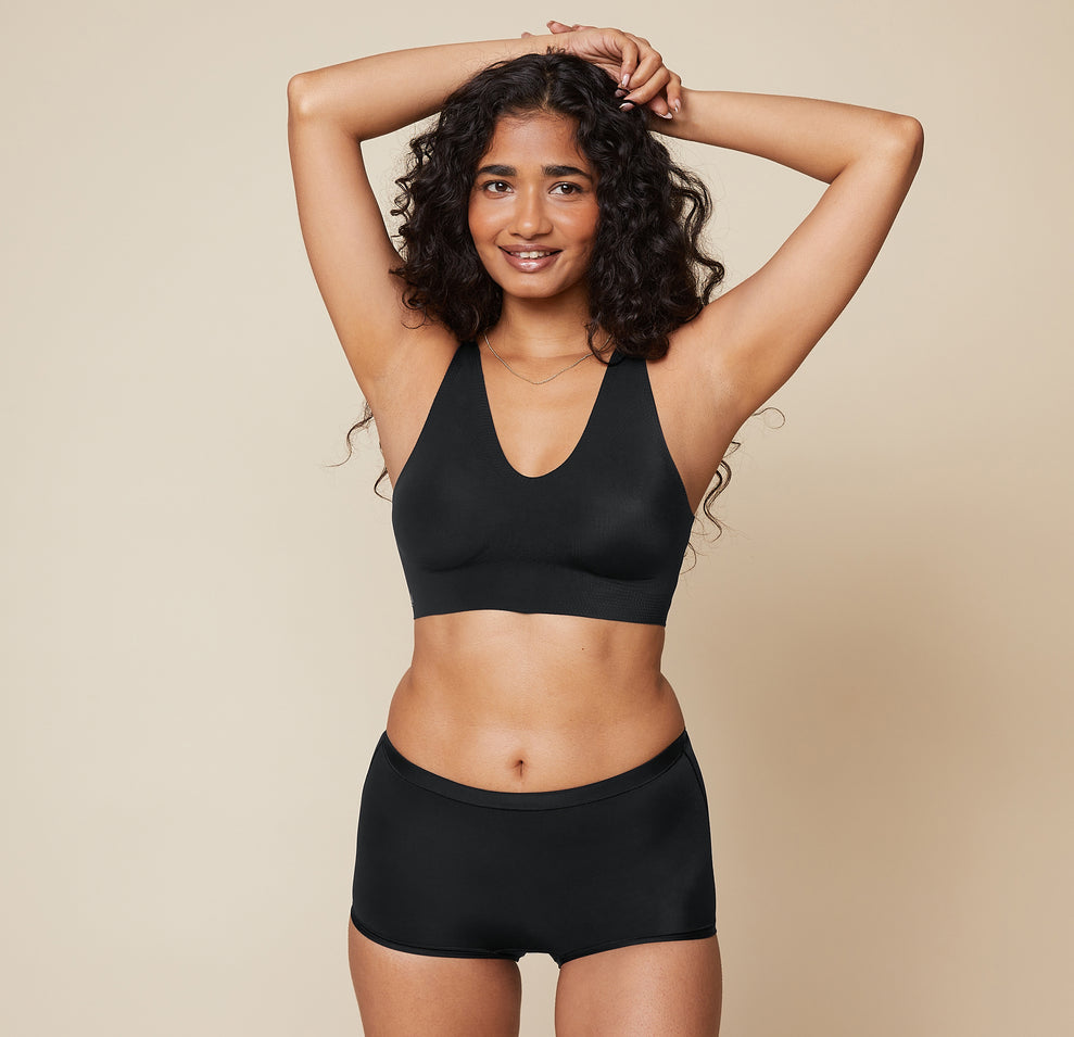 Naisha smiles with arms raised, wearing the Bliss Sleek Lined Bralette and Bliss Boyshort in Black. The bralette features a seamless design and V-neckline, paired with the comfortable boyshorts.