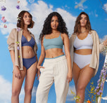 Carli, on the left, poses confidently wearing the Bliss Triangle and Bliss Hiphugger in Twilight. Naisha, in the center, wears the Bliss Scoop in Mist paired with light pants. Summerlyn, on the right, stands wearing the Bliss Bralette and Bliss Highwaist Brief in Tidal. All three models are set against a vibrant sky backdrop with delicate flowers in the foreground.