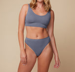 Megan stands facing forward, wearing the Cloud Cotton Scoop Bralette and Highwaist Thong in Slate. Her arms rest at her sides, and she gazes softly at the camera against a neutral background.