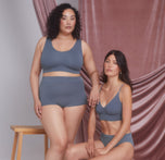 Two models sit together against a soft pink draped backdrop, one wearing the Cloud Cotton Scoop Bralette in Slate with the Highwaist Boyshort and the other wearing the Cloud Cotton Triangle Bralette in Slate. One model leans slightly on the other, creating a comfortable and relaxed atmosphere.