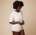 Model Akesha in the Cloud Cotton Tee in Heathered Oatmeal, standing in a casual, relaxed pose with a slight smile. The lightweight and breathable fabric is emphasized.