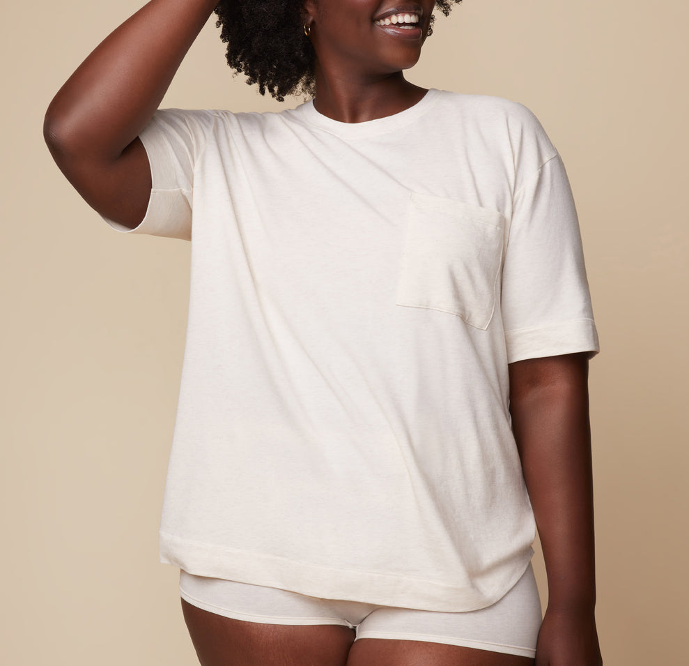 Model Akesha in the Cloud Cotton Tee in Heathered Oatmeal, with one hand resting on her head. The image showcases the relaxed fit and soft texture of the tee. sizing: Akesha wears size L