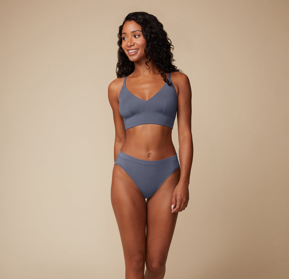Full-body view of the model smiling confidently, modeling the Cloud Cotton Triangle Bralette and matching bottom in Slate. She stands with one hand resting lightly on her hip.