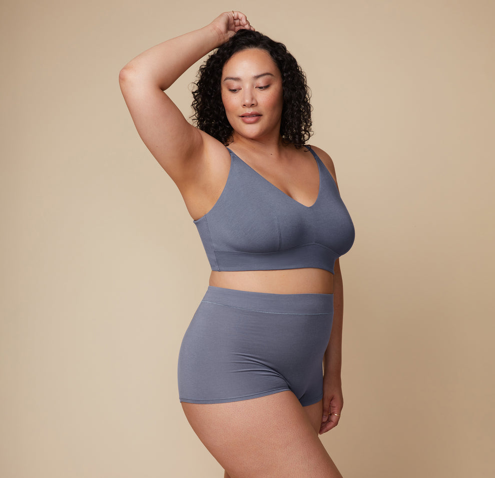Model poses with one hand resting on her head while wearing the Cloud Cotton Triangle Bralette and matching bottom in Slate. The neutral backdrop keeps the focus on her pose and the fit of the set.