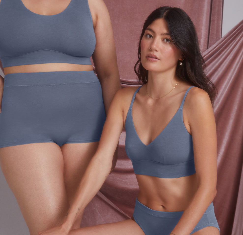 Two models sit side by side against a soft pink draped backdrop, Anna in the foreground wearing the Cloud Cotton Triangle Bralette in Slate. One model rests her arm gently on the other, creating a relaxed and comfortable scene.