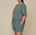 Model Summerlyn wearing the Pima PJ Shorts in Rainforest (Size: L) paired with the Base Bralette in Beige. The image emphasizes the elastic waistband and comfortable, casual style of the shorts.