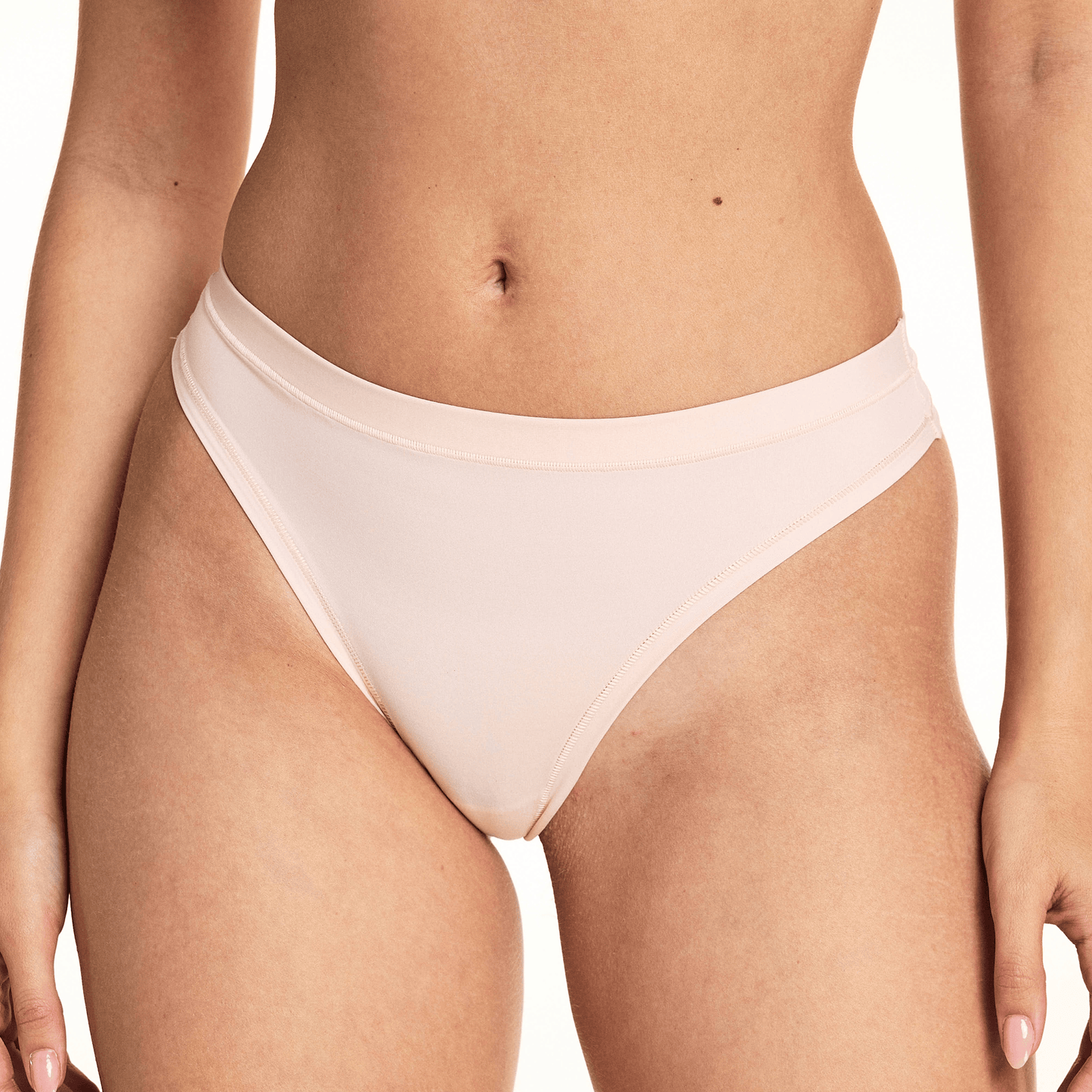 In a studio setting, Laura wears the Bliss Thong in Rose, featuring a smooth microfiber finish, low-rise silhouette, and minimal back coverage with soft elastic trim along the waistband and leg openings.