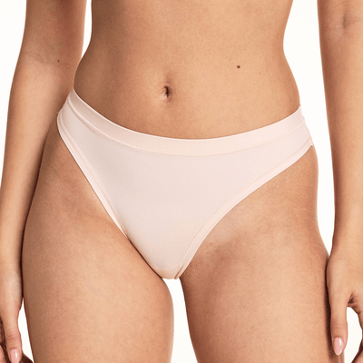 In a studio setting, Laura wears the Bliss Thong in Rose, featuring a smooth microfiber finish, low-rise silhouette, and minimal back coverage with soft elastic trim along the waistband and leg openings.
