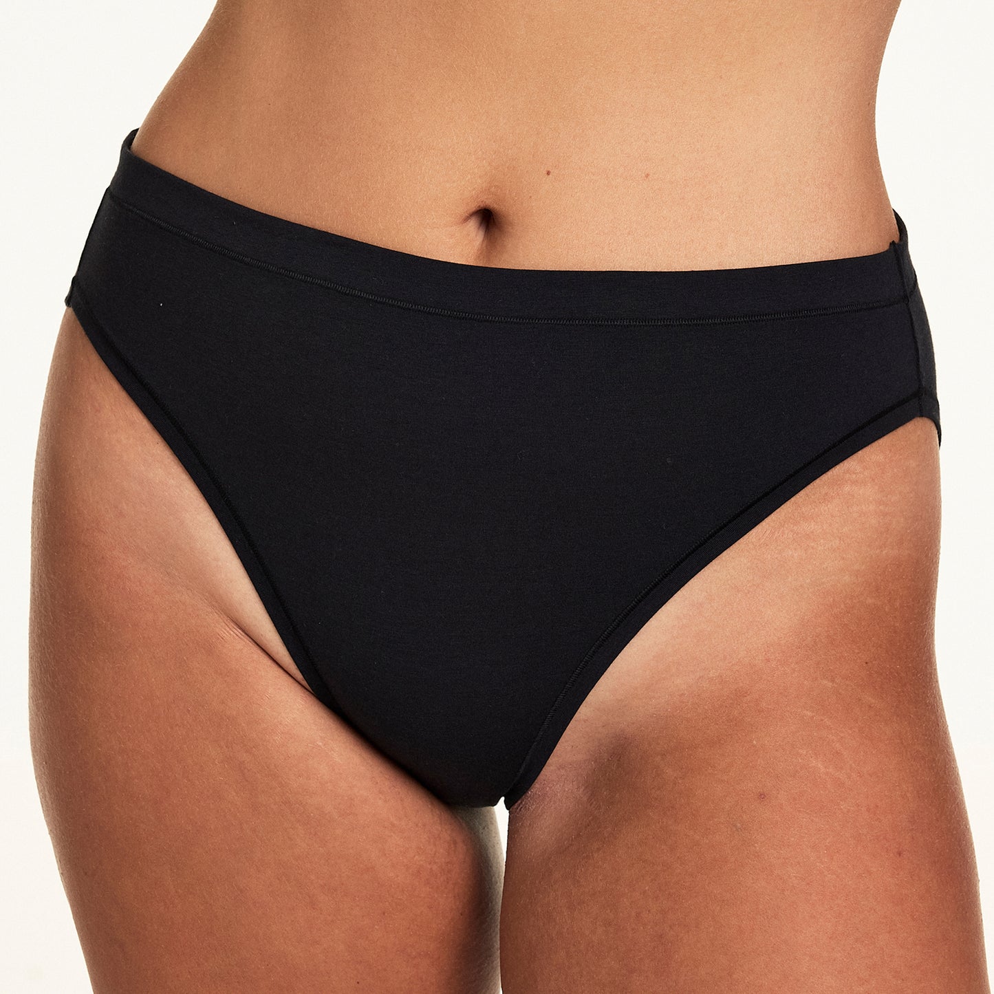Close-up on a light studio backdrop of Sonnie wearing the Cloud Cotton French Cut in Black with a matching black bra. The underwear features smooth cotton fabric and a high-rise waistband.