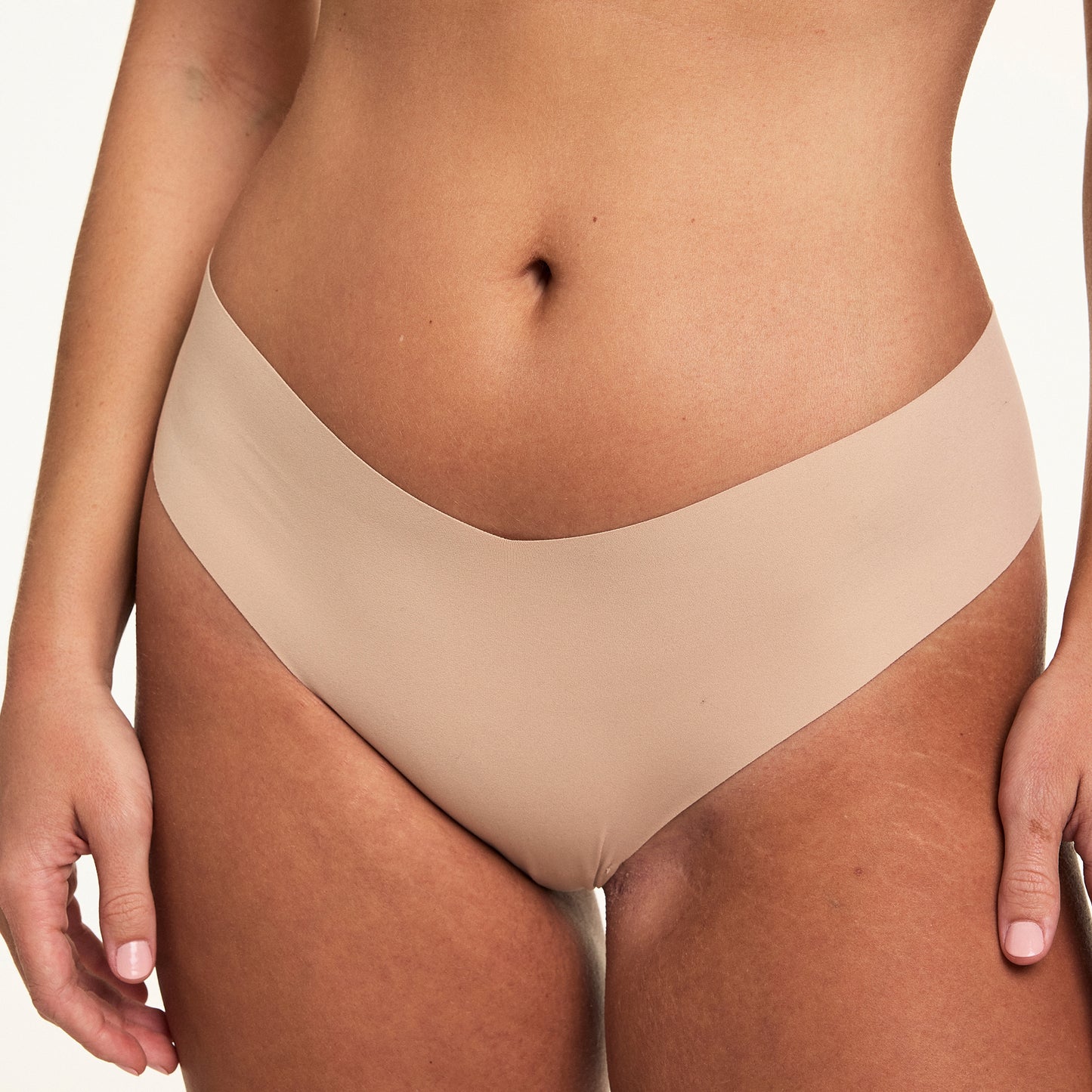 On a white studio backdrop, Sonnie wears the No-Show Thong in Beige, featuring a raw-edge, elastic-free design with a smooth, second-skin fit and low-rise silhouette.