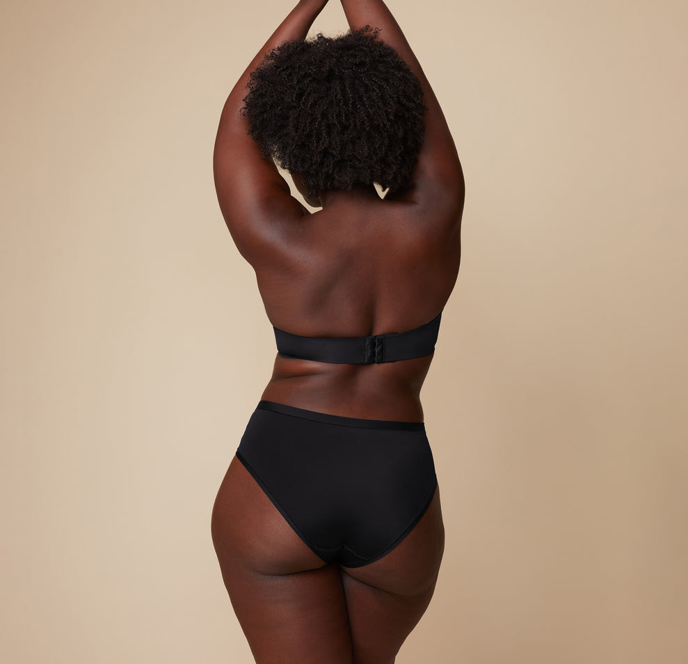 Back view of Akesha modeling the Base Strapless Bra in Black. Her arms are lifted, showing the strapless bra's back closure and band.