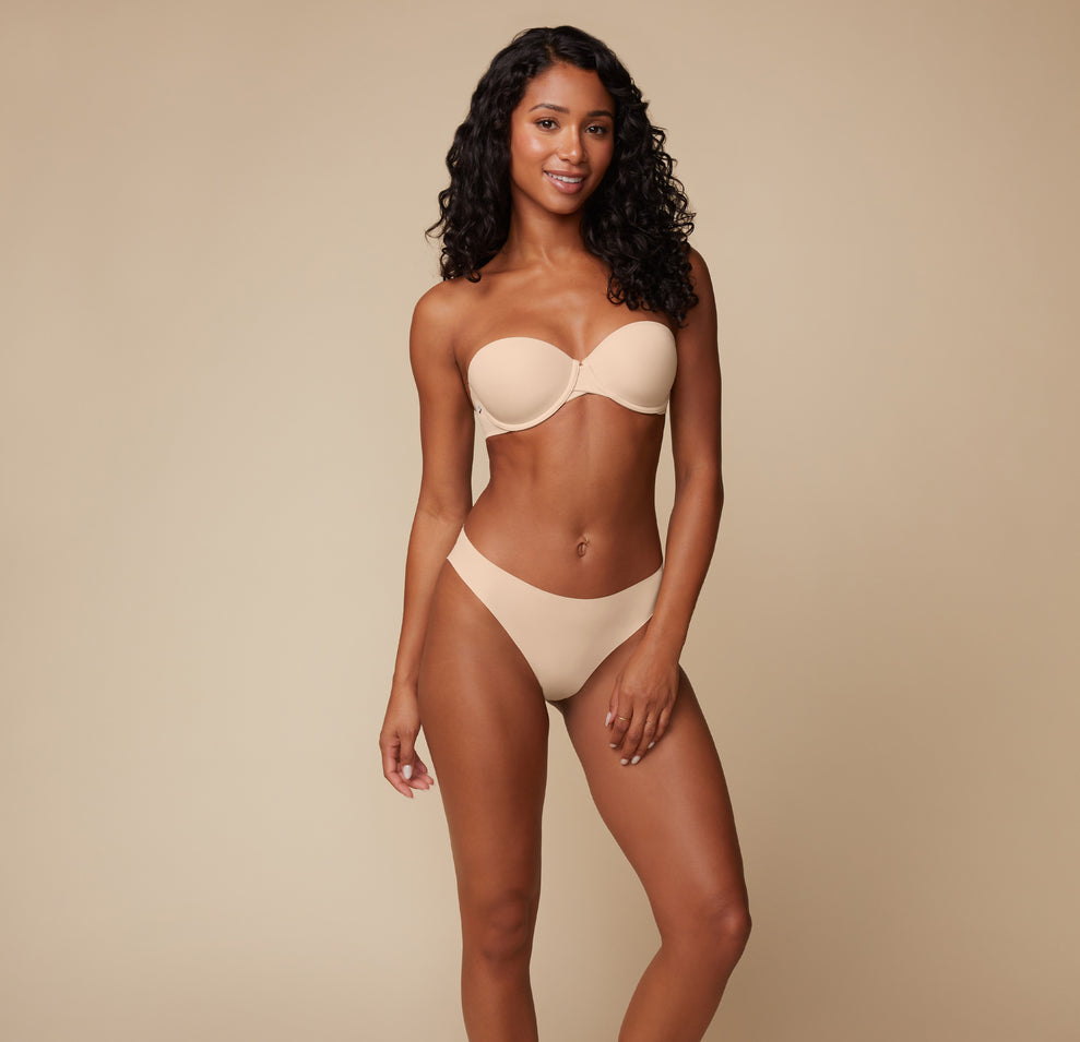 Ally stands confidently, wearing the Base Strapless Bra in Beige. She poses with one hand resting on her hip, showing the smooth and supportive design of the strapless bra.
