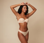 Full-body view of Ally posing with her hands behind her head, modeling the Base Strapless Bra in Beige against a simple, neutral background.
