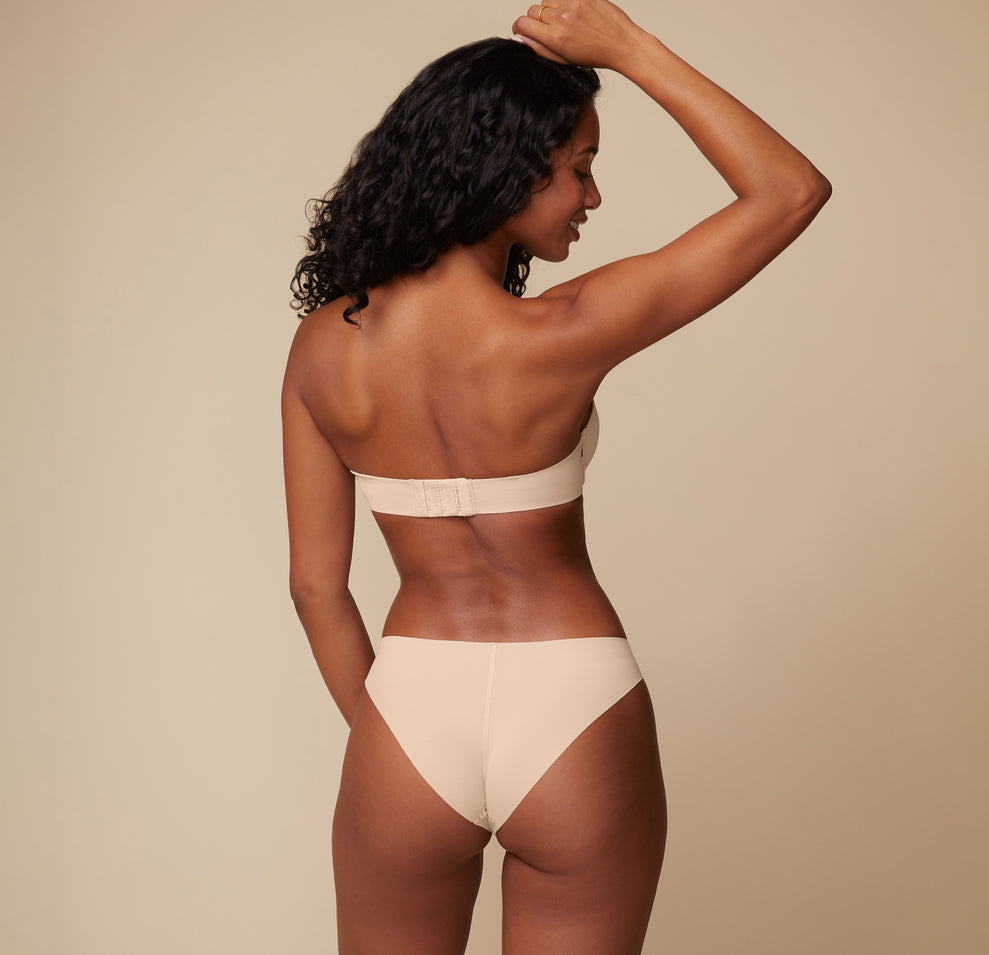 Back view of Ally modeling the Base Strapless Bra in Beige. She poses with her arms lifted, emphasizing the back closure and smooth band of the strapless bra.