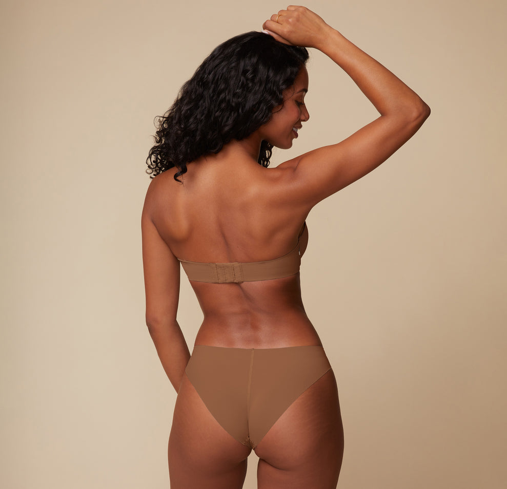 Back view of model Ally in the Base Strapless Bra in Brown, with arms lifted, displaying the wide back band for a secure, no-slip fit.