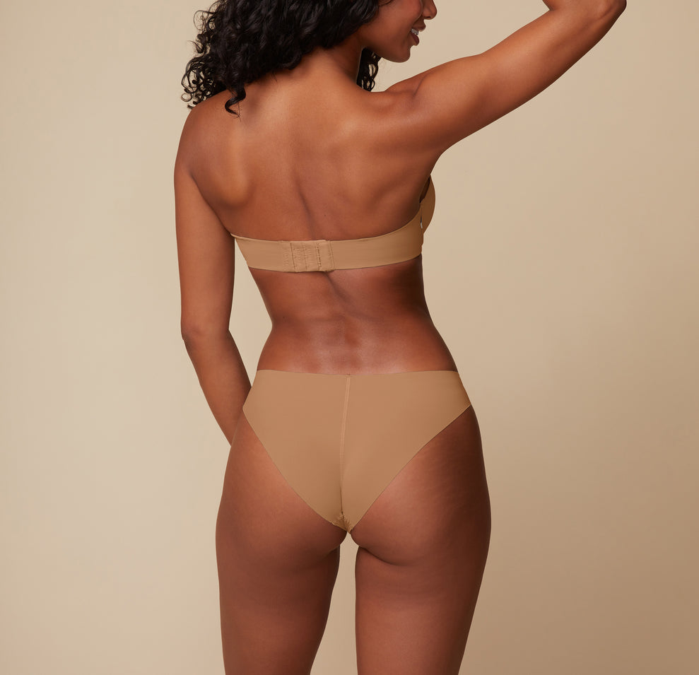A back view of Ally, showing the minimal coverage of the No-Show Bikini in Tan. Her arms are raised, drawing attention to the seamless design and comfortable fit.