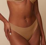 A close-up side view of Ally wearing the No-Show Bikini in Tan, with her left hand resting on her hip. The image focuses on the smooth waistband and sleek silhouette.