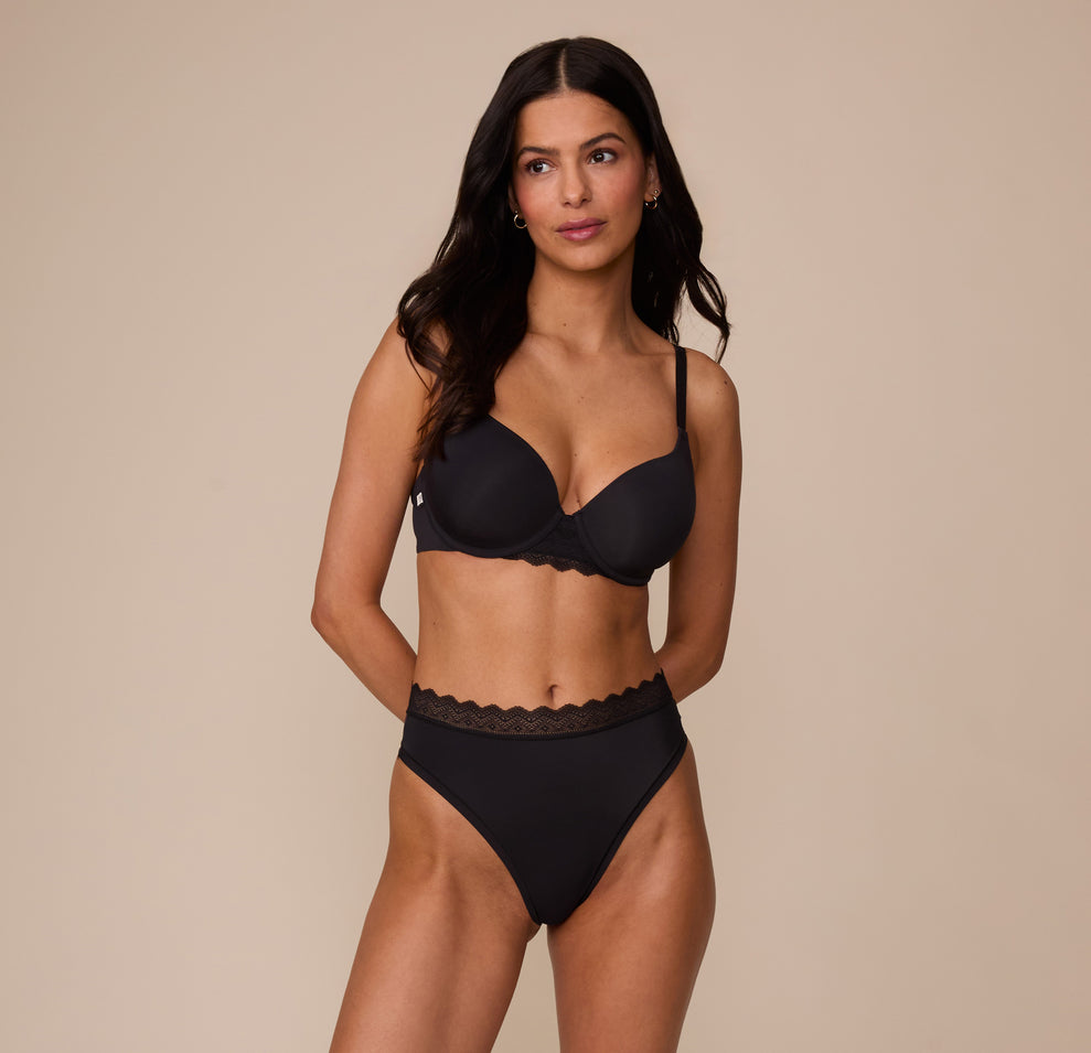 Marcela in a full-body pose wearing the Base Lace T-Shirt Bra and matching Bliss Lace Highwaist Thong in Black. The image emphasizes the elevated everyday design with soft lace trim and breathable fabric.