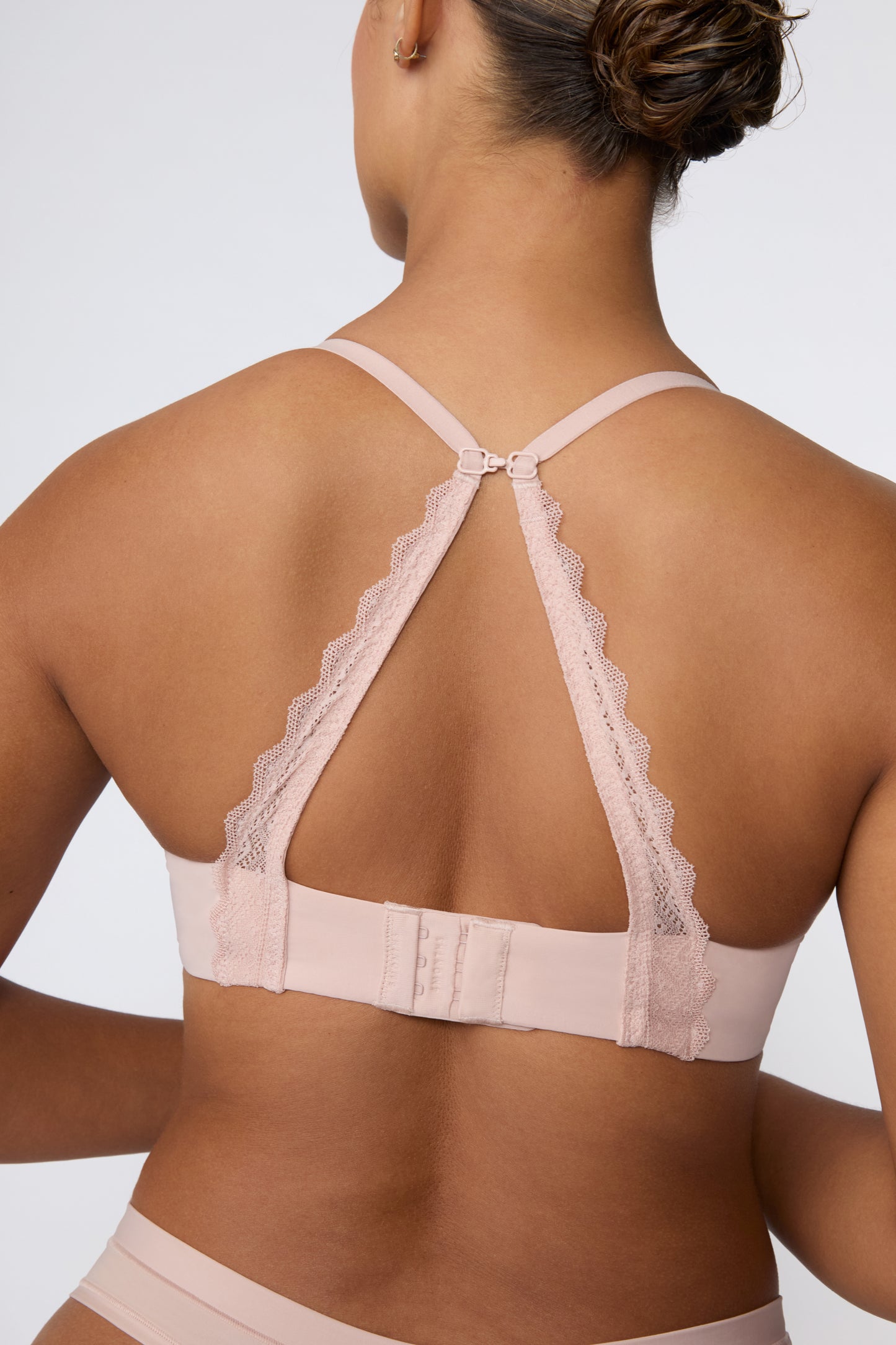 In a studio with a light gray backdrop, India wears the Base Lace Bra in Ember Rose from the back, showing the lace racerback conversion with sliding J-hook and hook-and-eye closure.