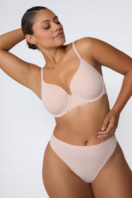 In a studio with a light gray backdrop, India wears the Base Lace Bra in Ember Rose with matching underwear, showing the lightly lined cups, lace trim along the band, and smooth microfiber fabric.