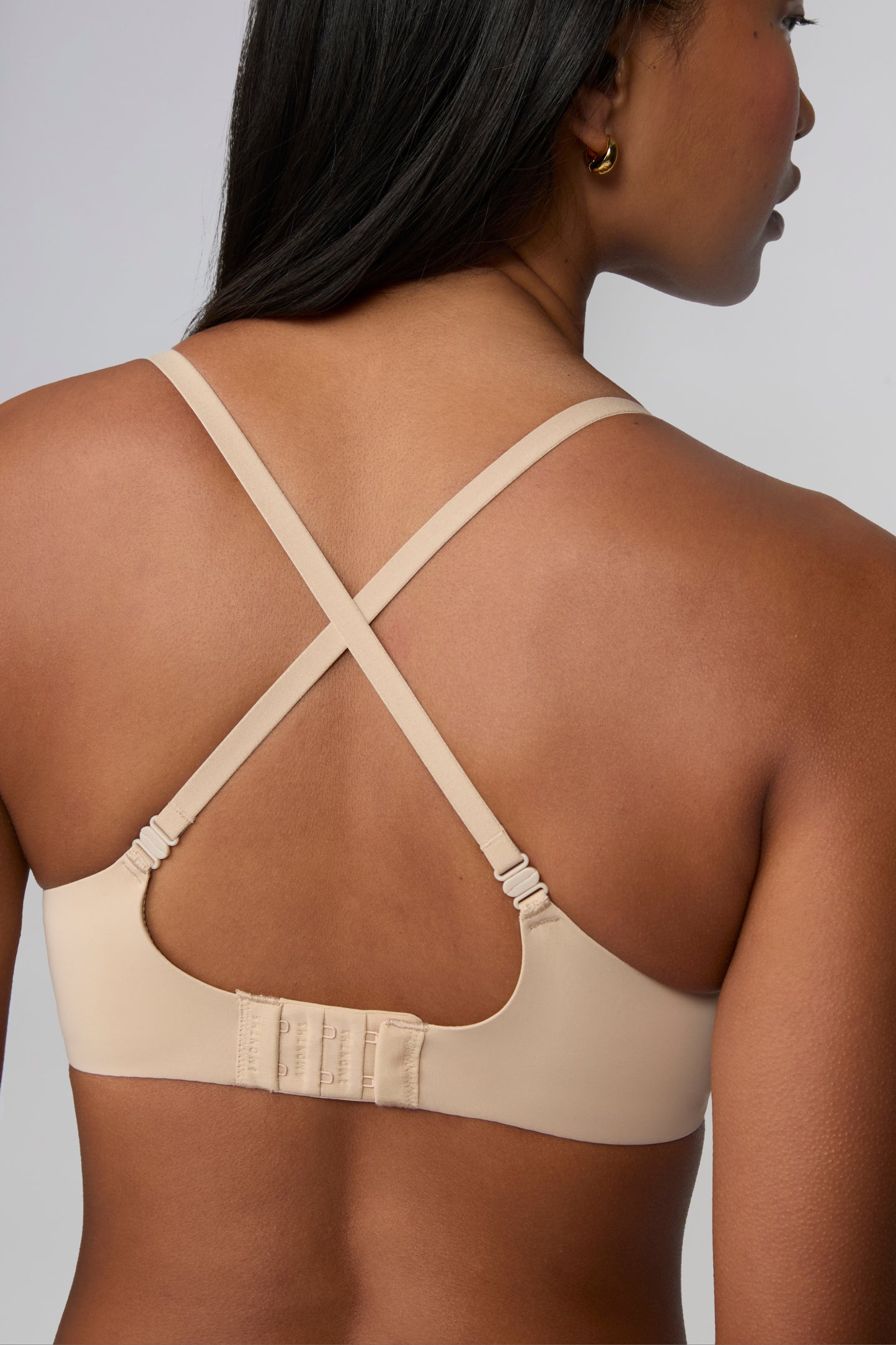 On a light grey studio backdrop, Amber wears the Base Push-Up Bra in Beige, shown from the back with straps crossed. The convertible straps form an X shape, and the smooth microfiber band features a hook-and-eye closure.