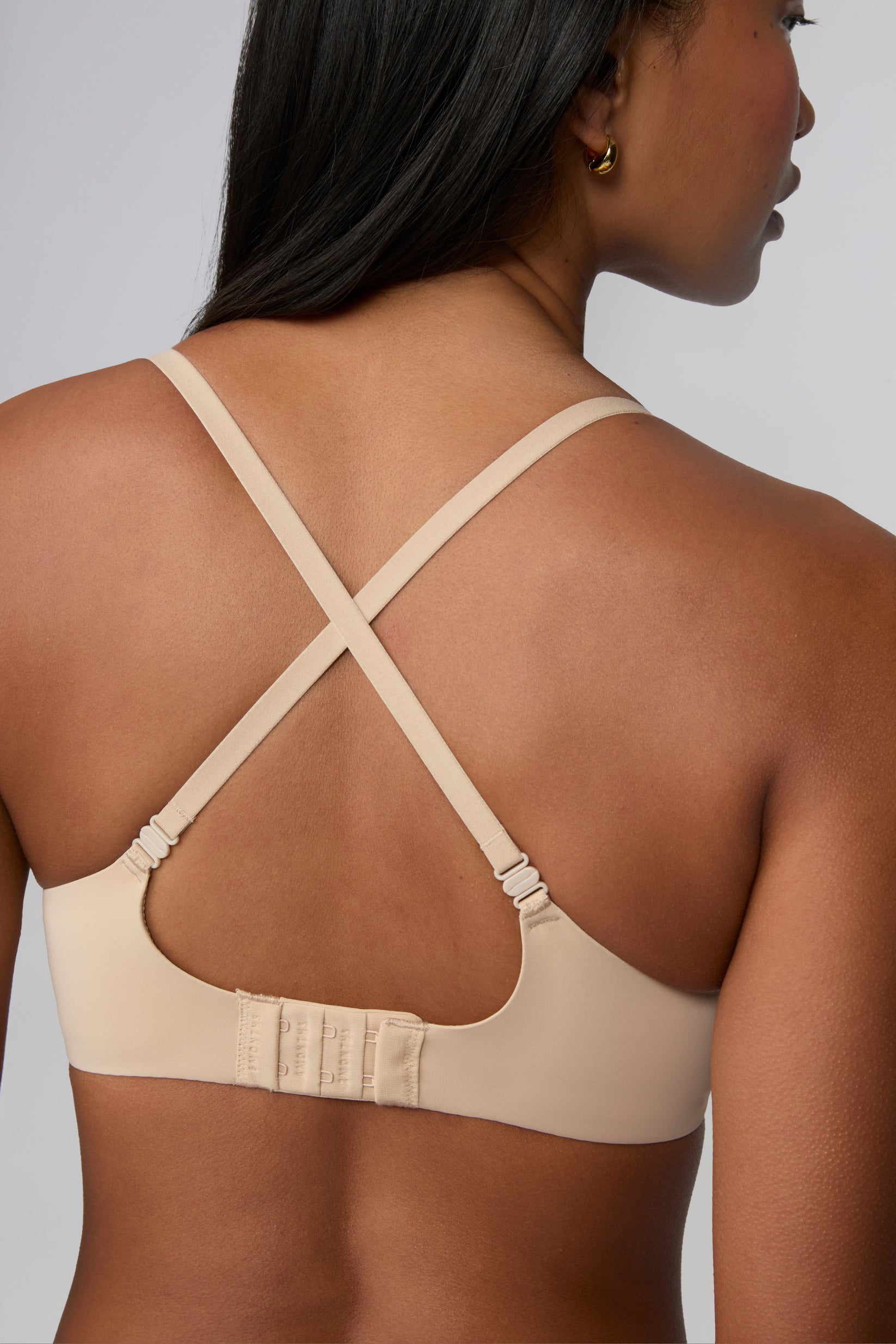 On a light grey studio backdrop, Amber wears the Base Push-Up Bra in Beige, shown from the back with straps crossed. The convertible straps form an X shape, and the smooth microfiber band features a hook-and-eye closure.