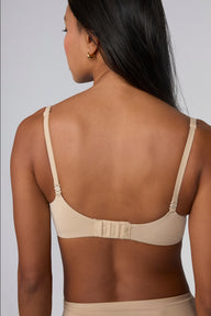 Against a light grey background, Amber models the Base Push-Up Bra in Beige from the back with straps worn straight. The adjustable straps and hook-and-eye closure are visible, with the soft microfiber band laying smooth across the back.